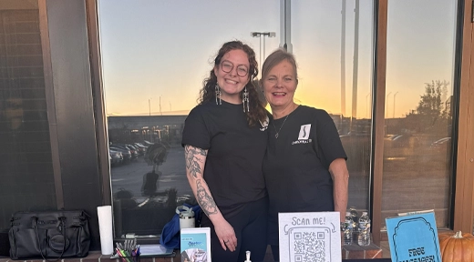 Dr. Pamela Fernandes and staff member stand behind a table with a QR code sign, water bottles, and items, in front of a glass window at sunset.